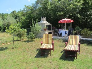 Due sedie a righe da giardino, un barbecue in pietra e un ombrello rosso in un ambiente naturale.