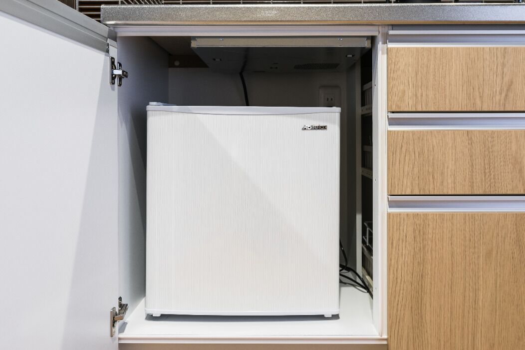 Een compacte, witte koelkast met een logo, in een keuken met houten kastjes en een metalen aanrecht.
