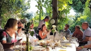 A group of people dining at an outdoor table with a white tablecloth, surrounded by greenery.