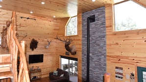 Interior de una cabaña de troncos con paredes de madera, una chimenea de piedra, cuernos de ciervo colgados y un televisor.