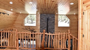 Interior de una cabaña con paredes de madera, chimenea de piedra y baranda de madera.