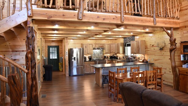 Interior rústico de una cabaña con escalera de madera, cocina con electrodomésticos modernos, zona de comedor con muebles de madera y habitación superior.