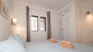 A bedroom with a bed, two windows with curtains, a wall-mounted lamp, and a door with louvered shutters. - VM7, first floor. (El Cotillo)