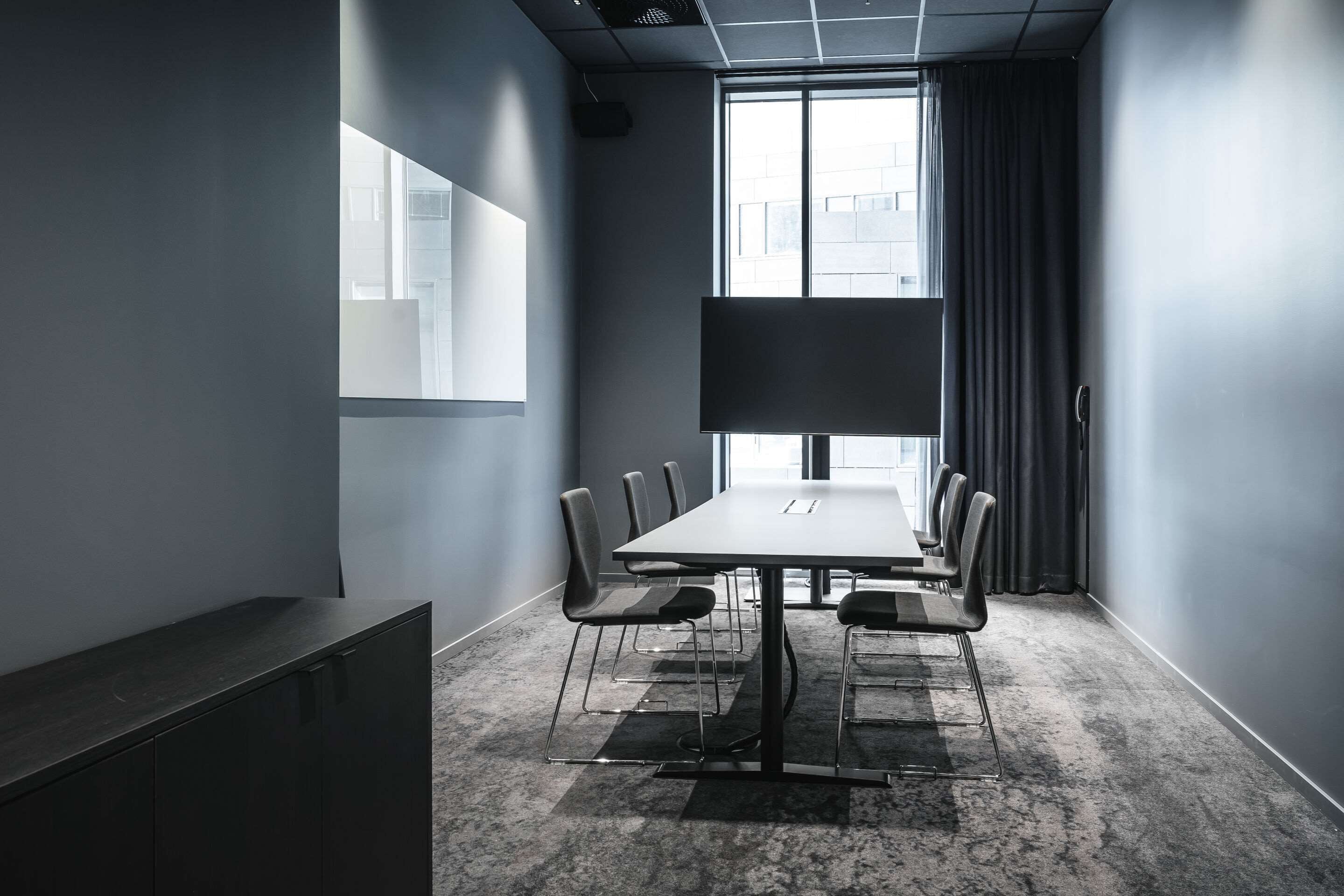 Une salle de conférence avec une longue table, six chaises, un grand écran et une fenêtre avec des rideaux.