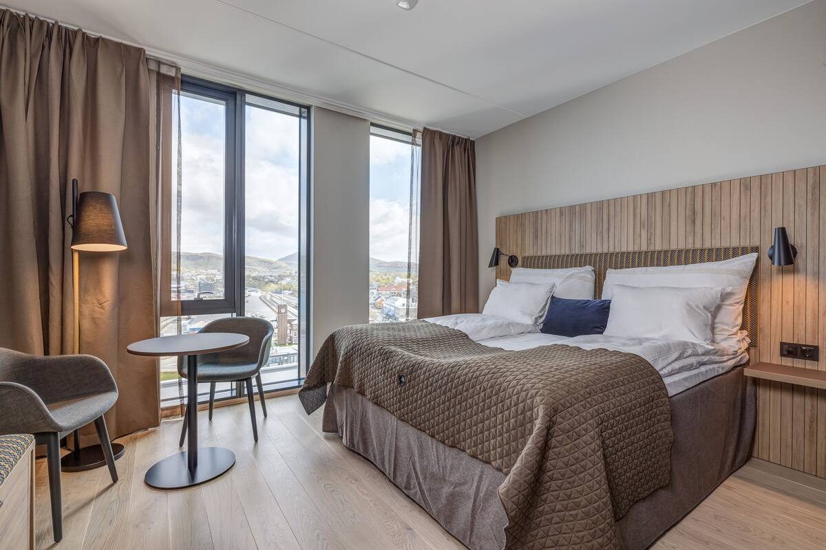 A hotel room with a large bed, a round table, and a chair. There is a view of the city through the window.