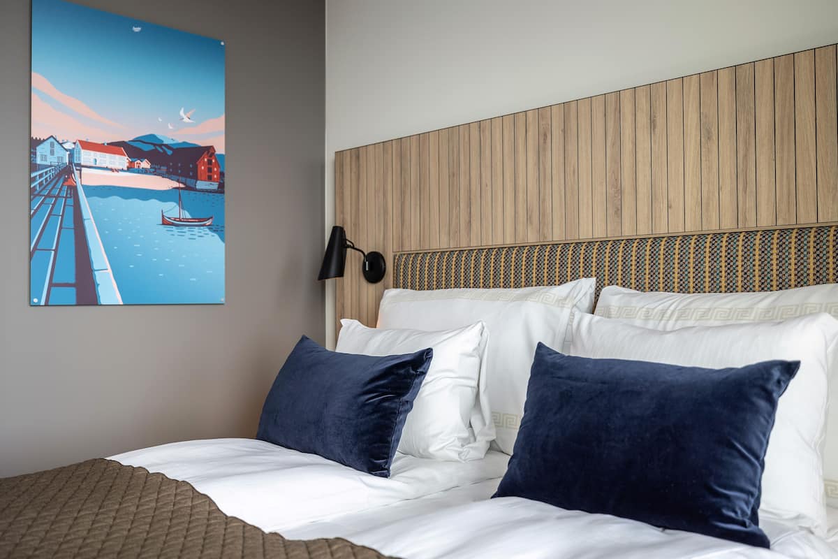 A hotel room with a bed, pillows, a wall-mounted lamp, and a painting of a coastal scene.