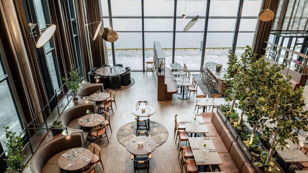 A modern restaurant with round tables and booths, large windows overlooking a body of water, and indoor plants. - Quality Hotel Ramsalt (Bodo)