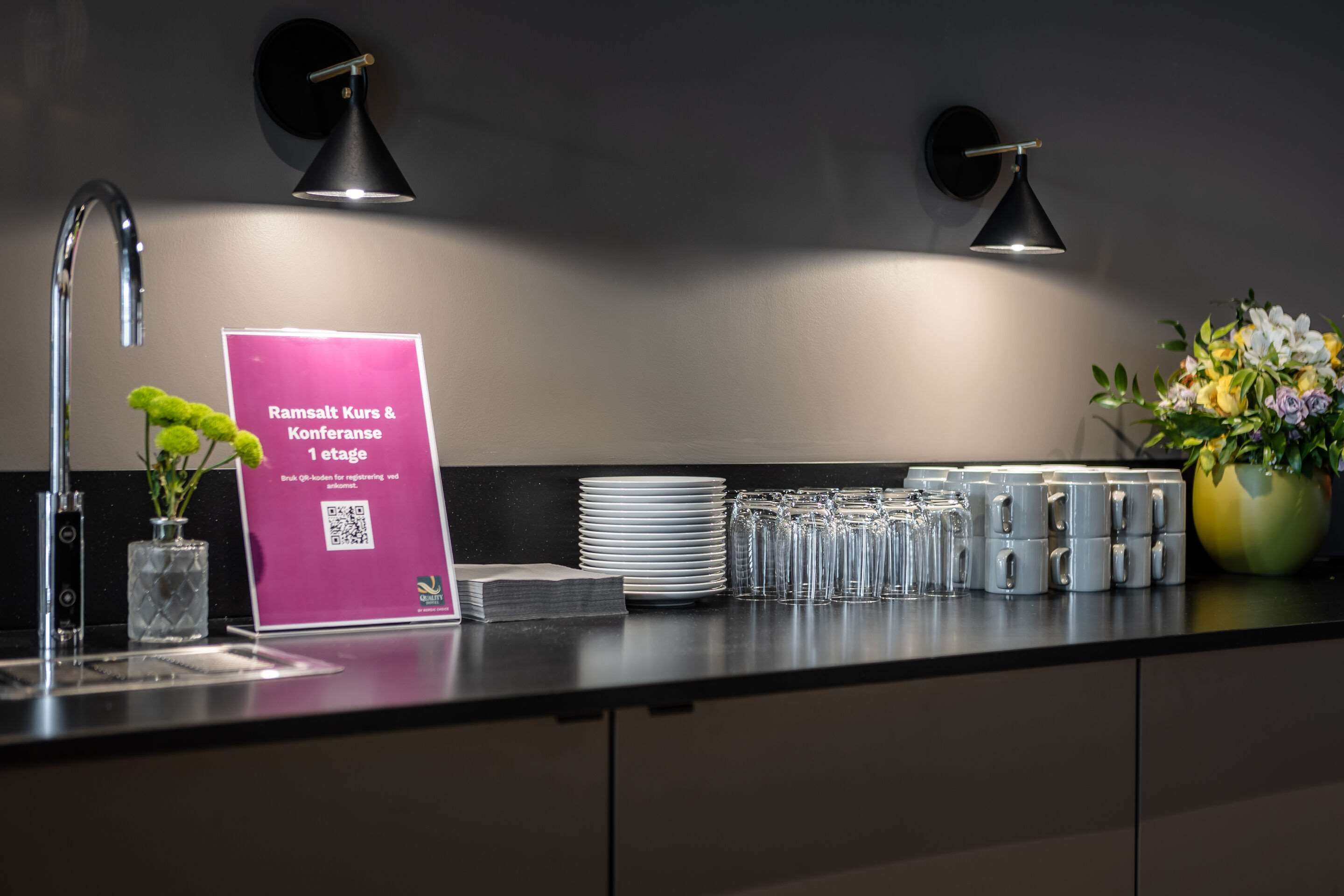 A modern kitchen counter with a sink, a QR code sign, a vase with flowers, and a stack of plates and glasses.