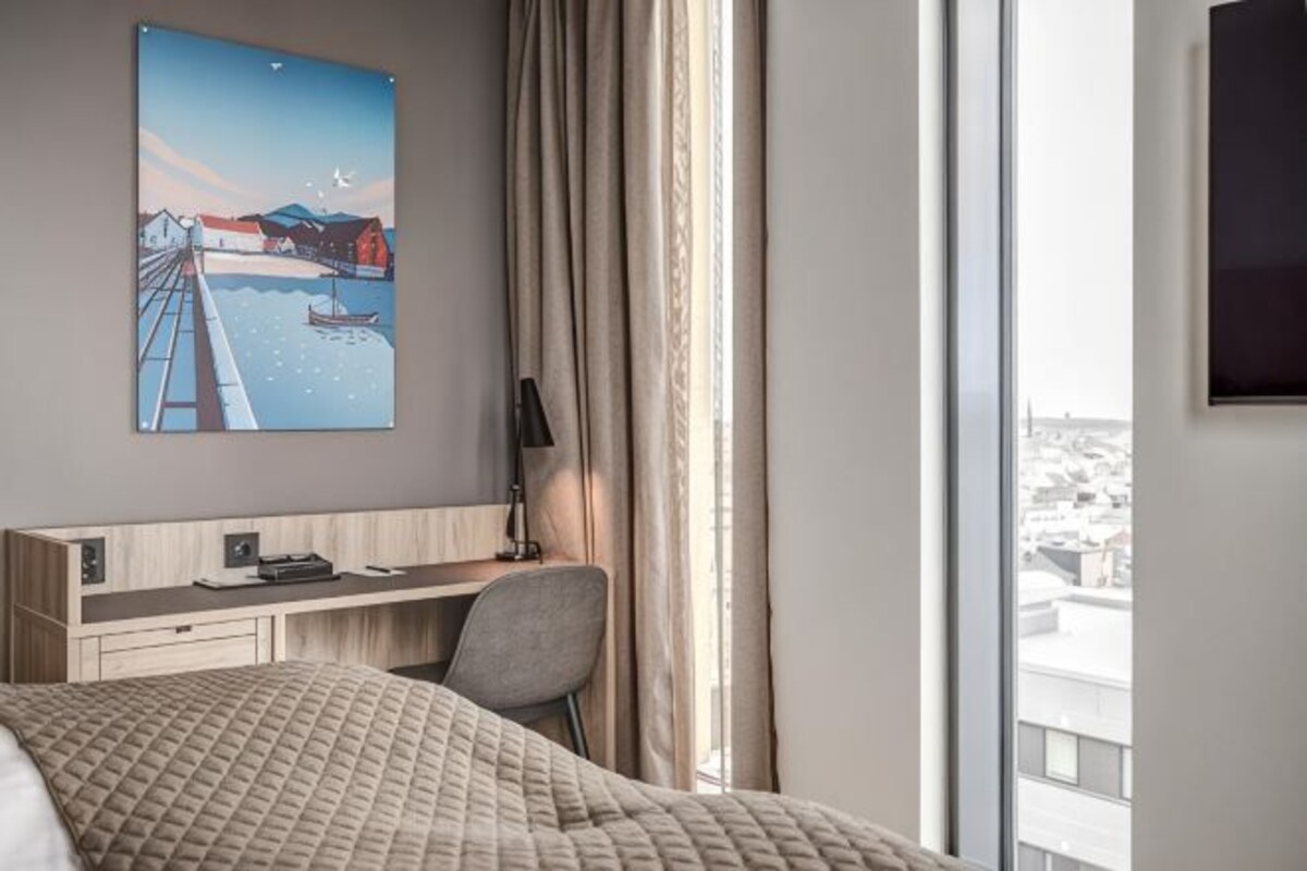 Ett hotellrum med en säng, ett skrivbord med en stol, en tavla, en tv och ett fönster med gardiner.