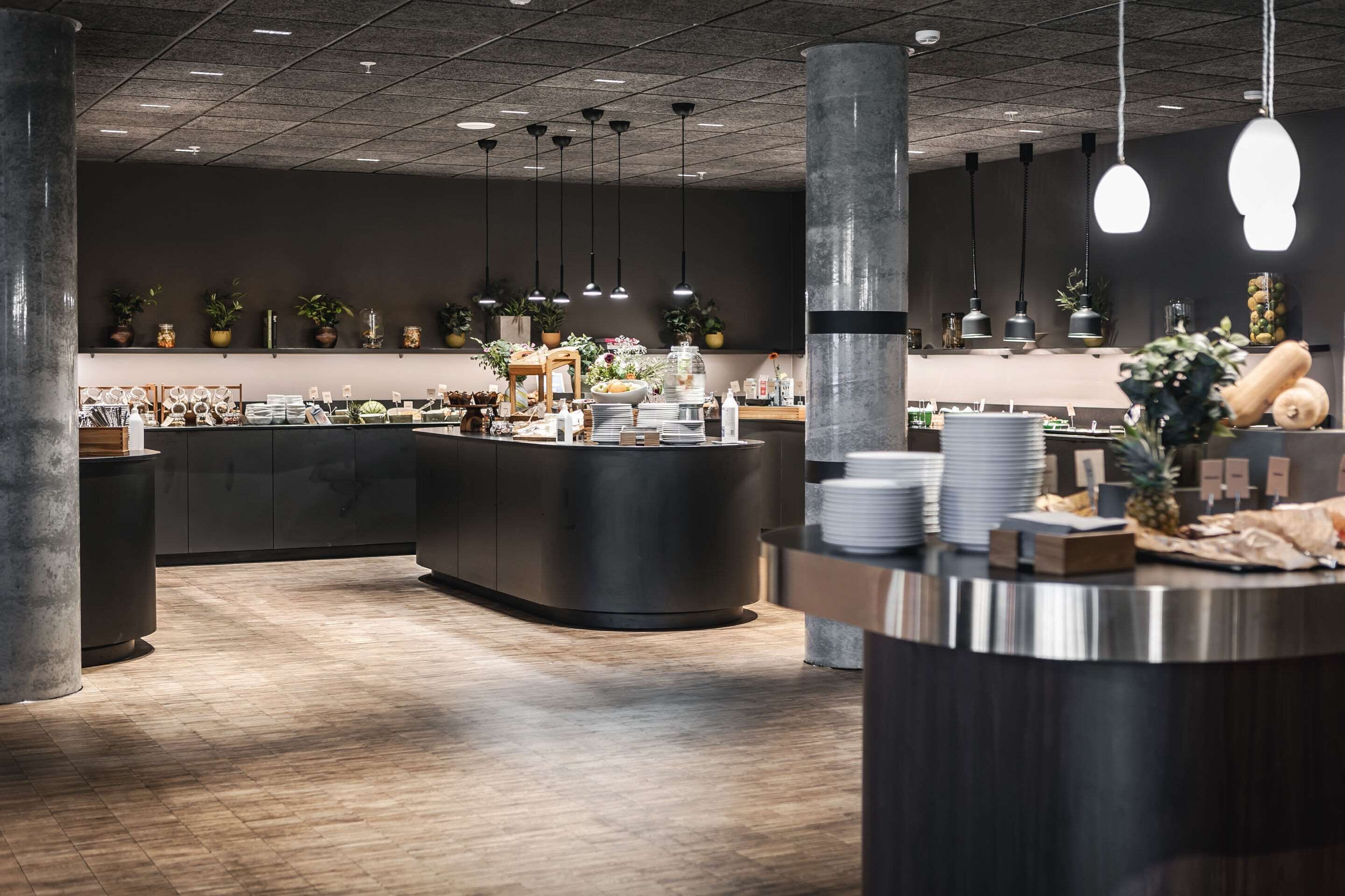 Een modern restaurant met een centrale balie, hangende lampen en potplanten.
