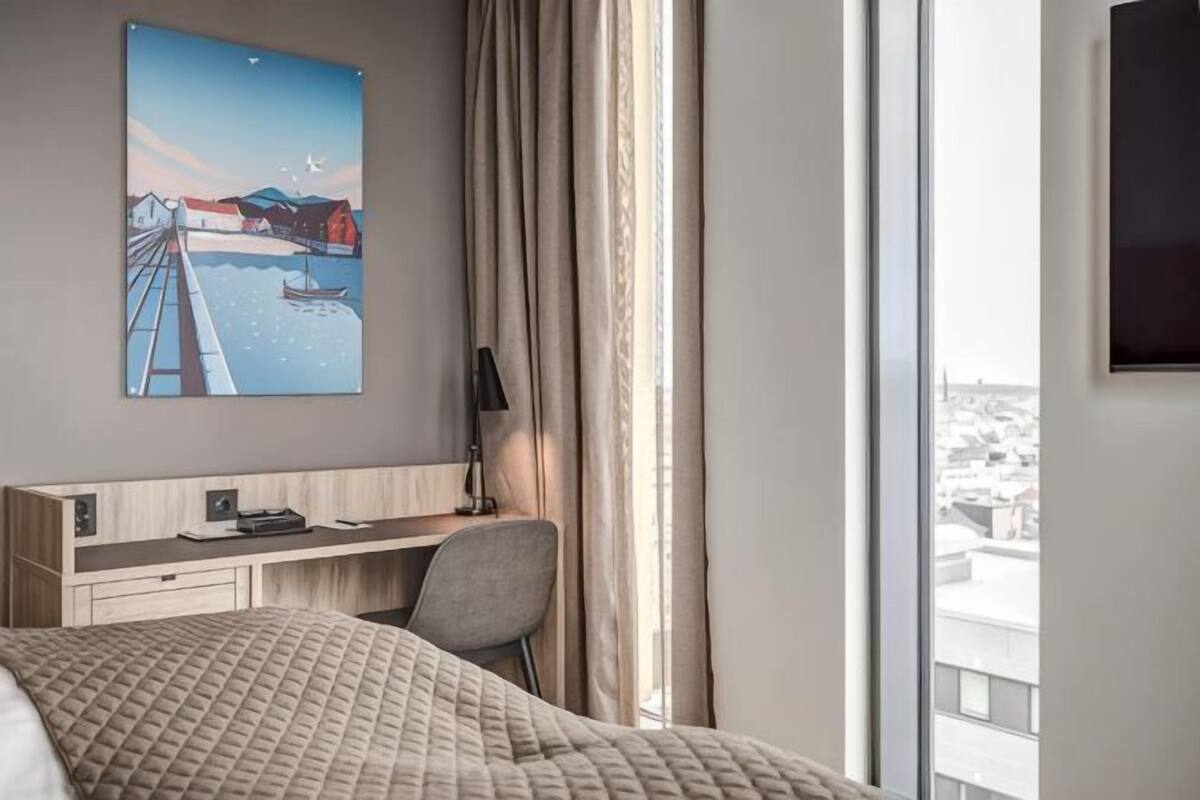 A hotel room with a bed, a desk with a chair, a painting, a TV, and a window with curtains.