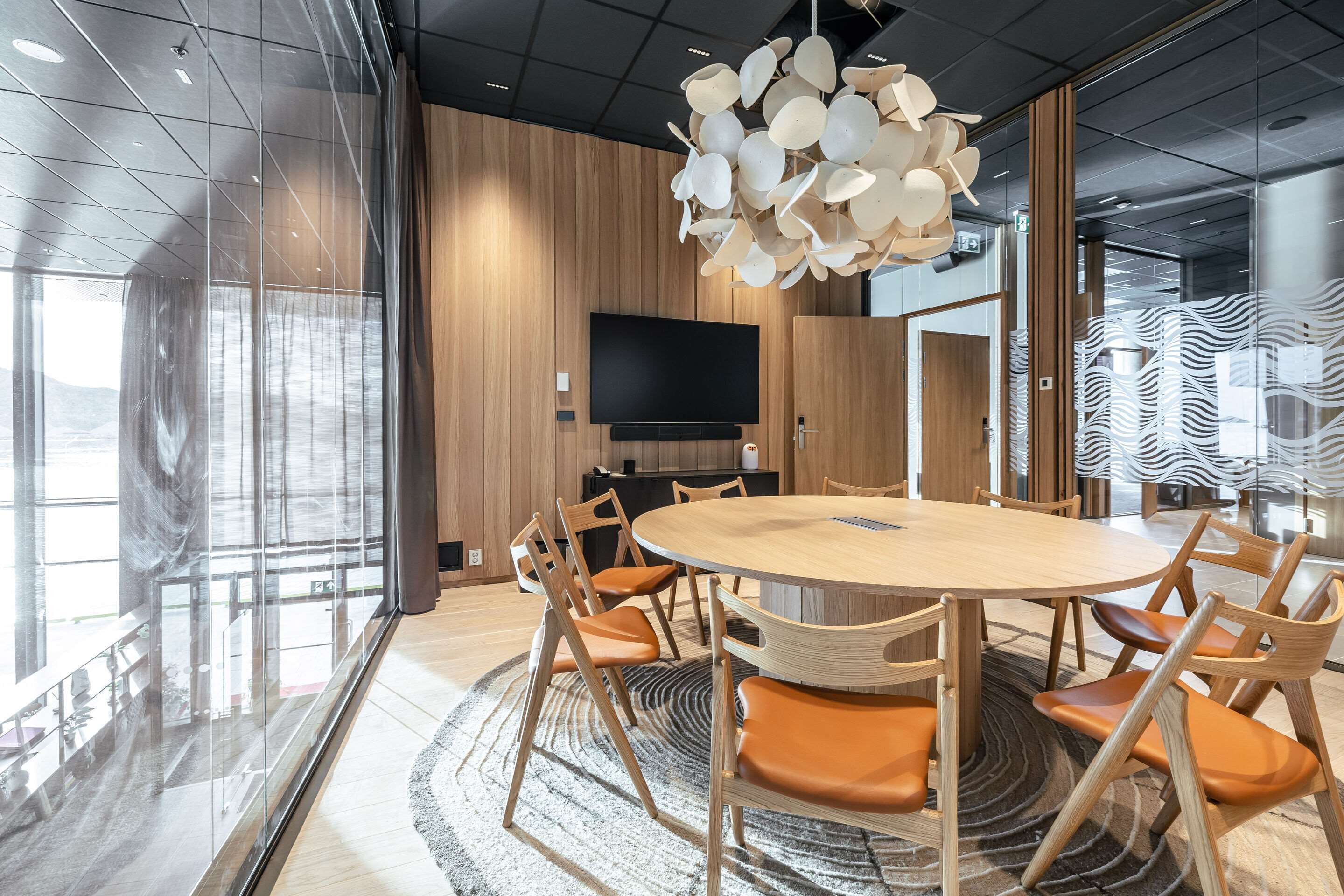 A modern conference room with a round table, wooden chairs, a large TV, and a unique ceiling light fixture.