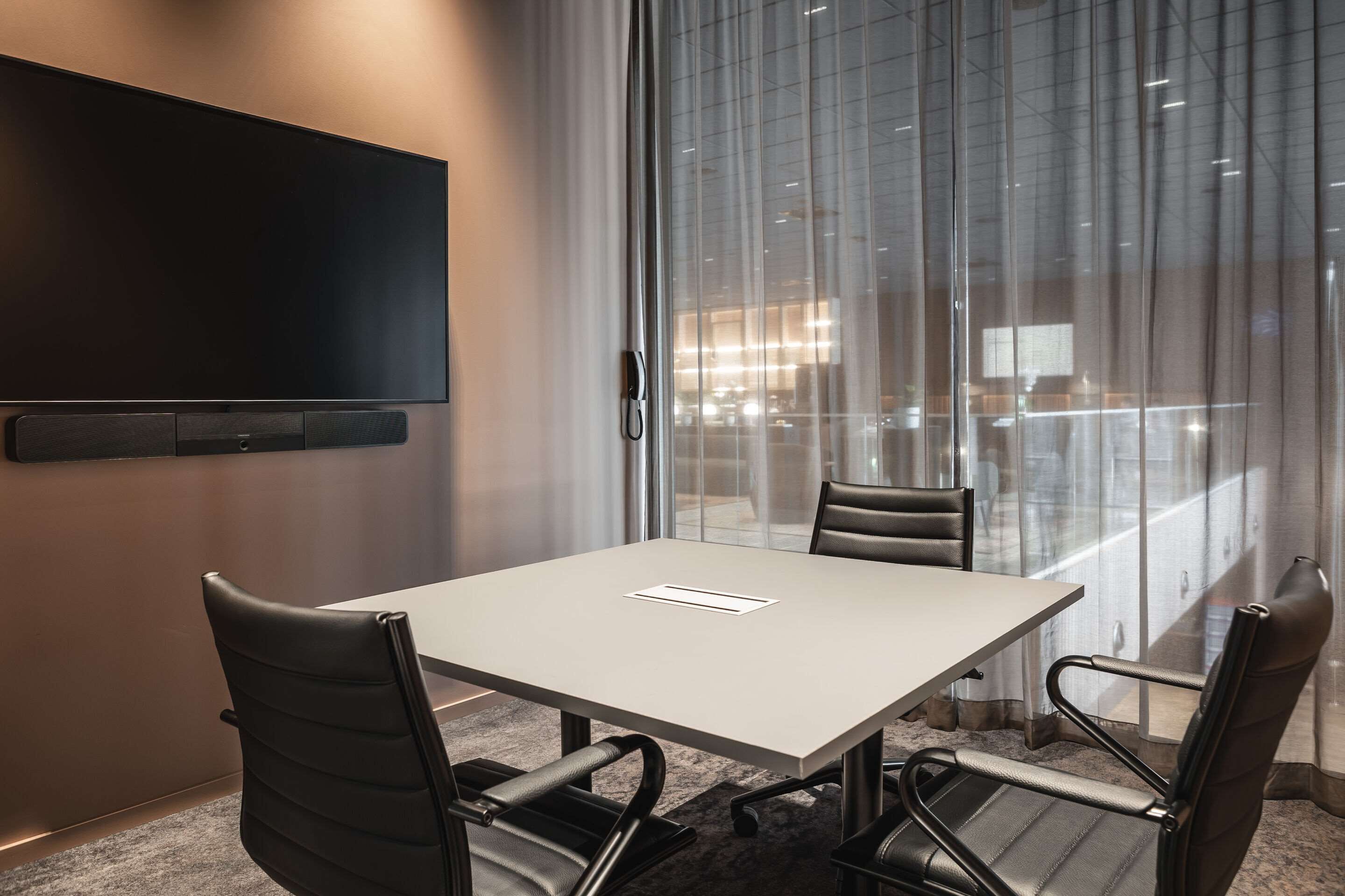 A conference room with a white table, four leather chairs, a flat-screen TV mounted on the wall, and large windows with sheer curtains.