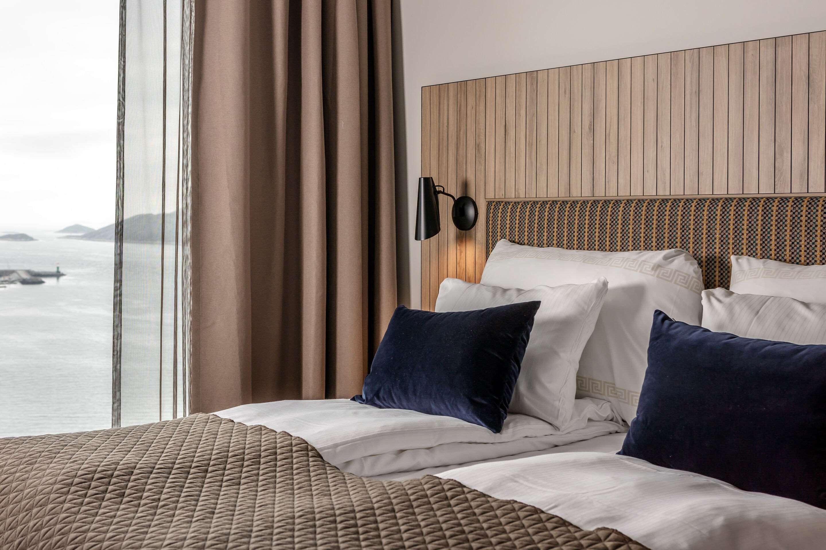 Habitación de hotel con una cama grande, cabecera con diseño de panel de madera, lámpara fijada a la pared y vistas al mar.