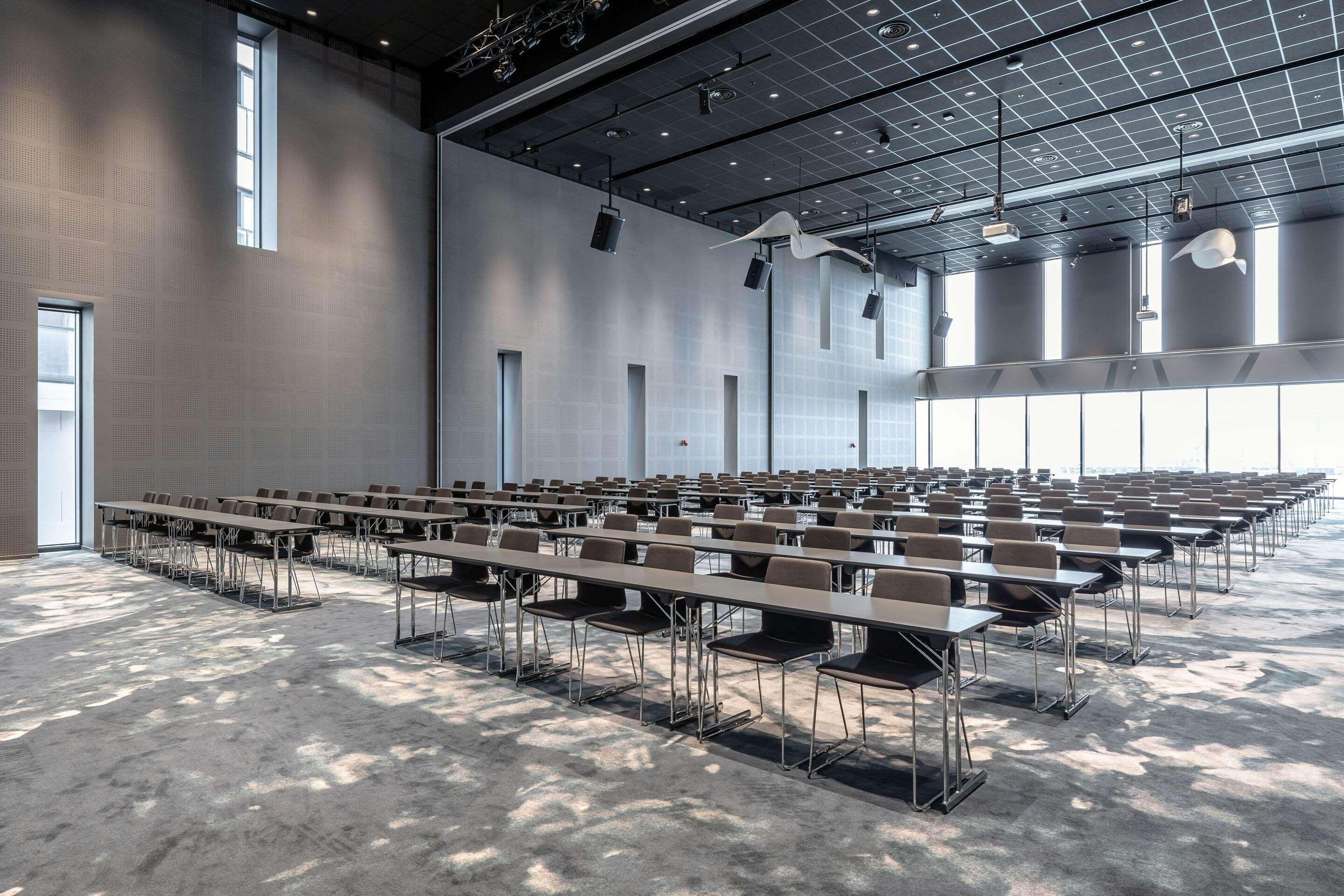 Un amplio salón de conferencias con filas de mesas y sillas, techos altos con patrones de cuadrícula y grandes ventanales que dejan entrar luz natural.