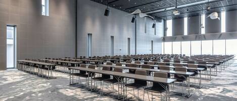 Un amplio salón de conferencias con filas de mesas y sillas, techos altos con patrones de cuadrícula y grandes ventanales que dejan entrar luz natural.