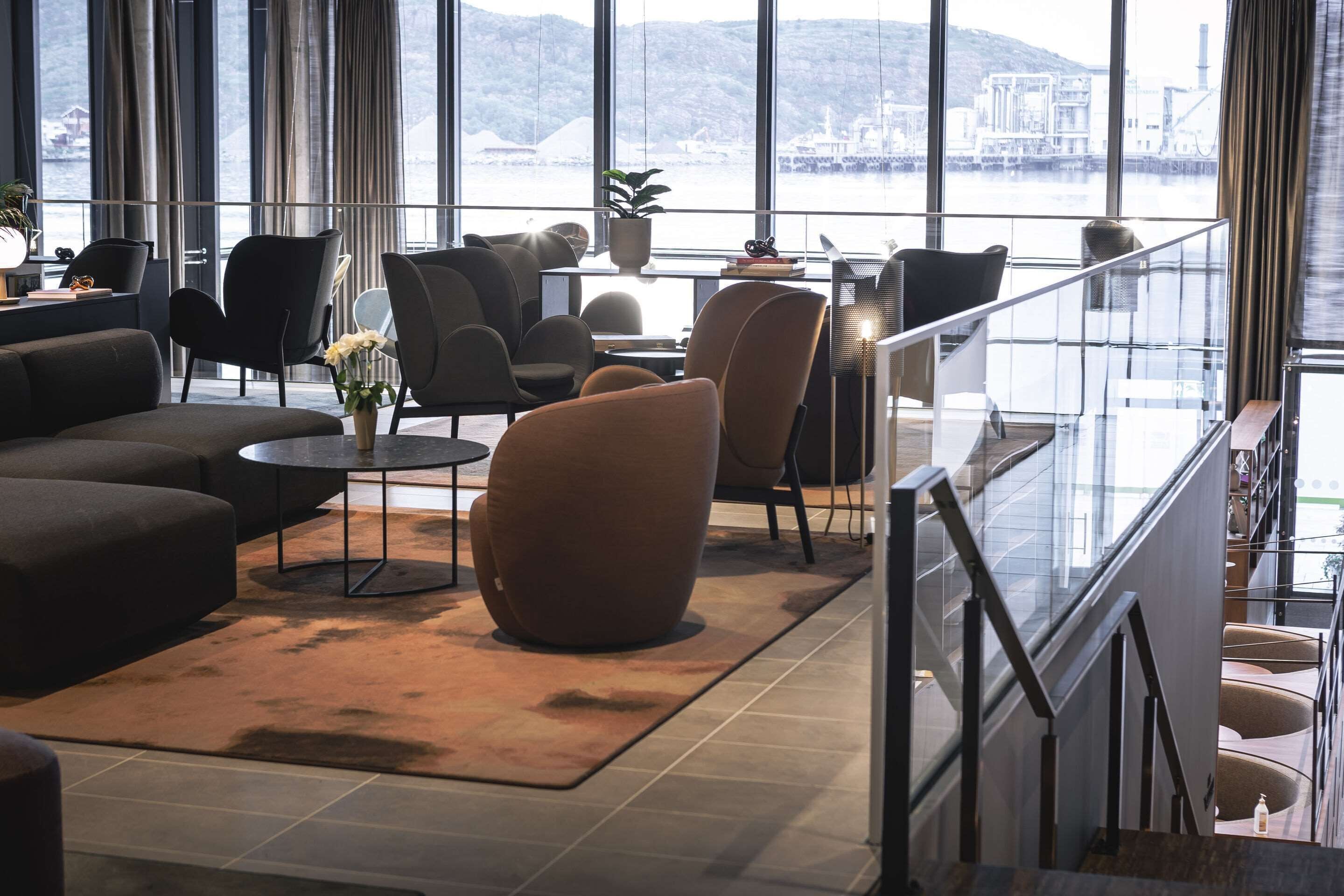 A modern hotel lobby with a large sofa, armchairs, a coffee table, and a view of a body of water through large windows.