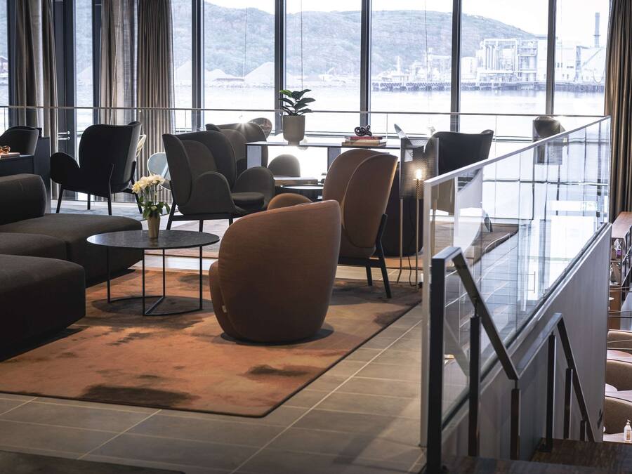 A modern hotel lobby with a large sofa, armchairs, a coffee table, and a view of a body of water through large windows.