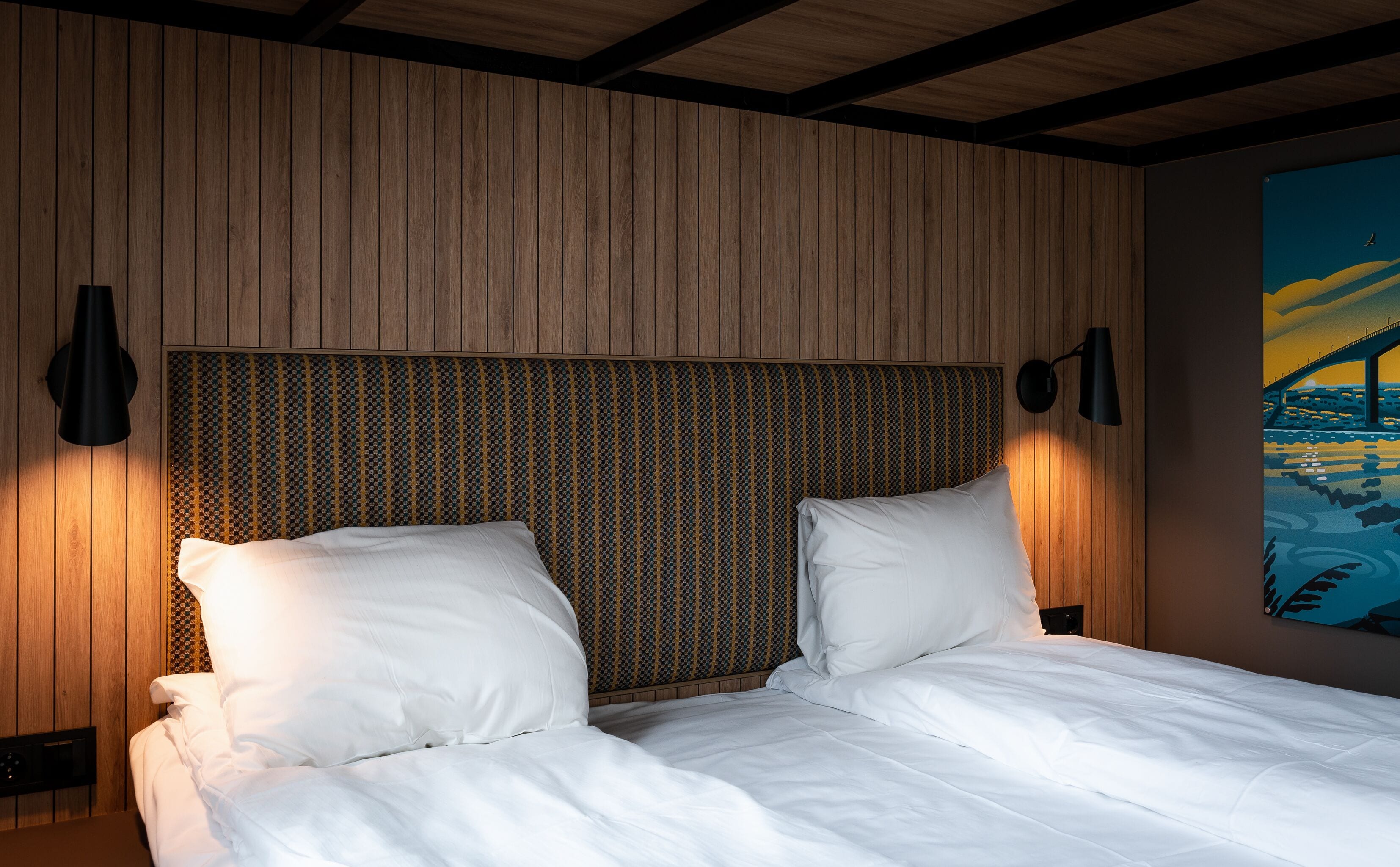 Un dormitorio con cabecera de madera, dos almohadas blancas y una mesita de noche con lámpara.