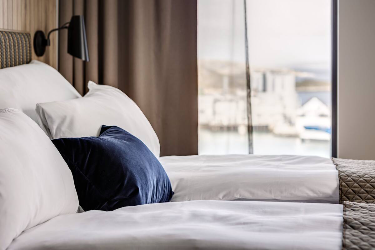 A hotel room with a bed, a view of boats through the window, and a lamp on the wall.