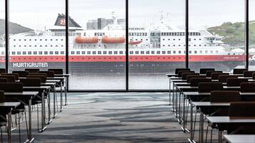 Ett konferensrum med rader av bord och stolar, stora fönster med utsikt över ett fartyg som heter ”HURTIGRUTEN”, som ligger förtöjt vid en hamn.