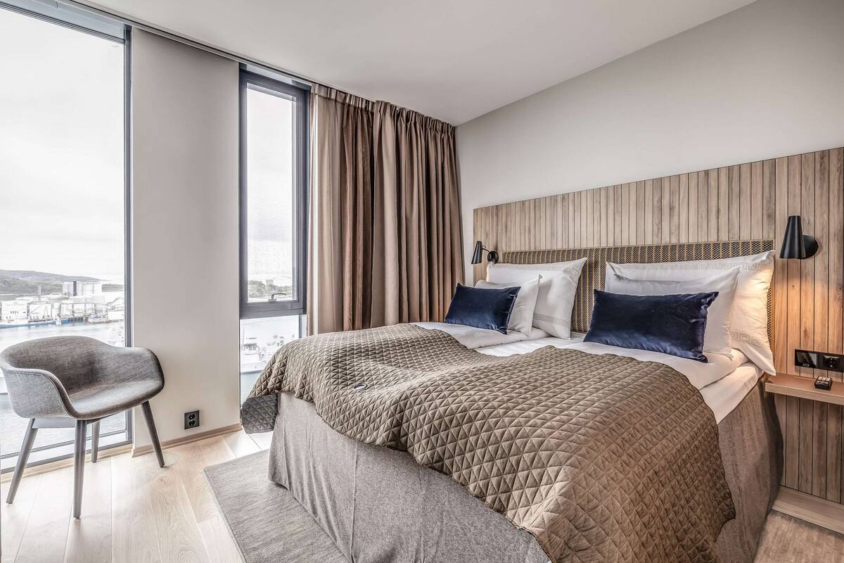 A modern hotel room with a large bed, a chair, a nightstand, and a view of the city.
