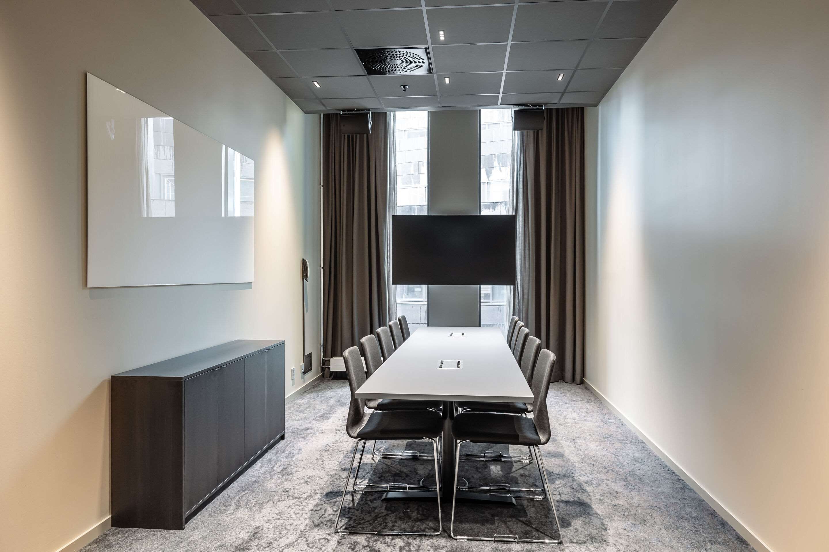 Une salle de conférence avec une longue table, des chaises, un buffet sombre, un grand écran et des rideaux.
