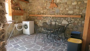 A stone-floored room with a washing machine, a table, chairs, and a trash bin.
