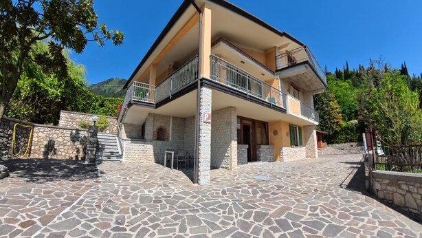 A two-story building with a stone-paved courtyard, a balcony with a metal railing, and a stone wall with a gate.
