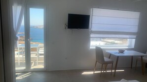 A room with a bed, a TV, a table with chairs, and a view of the sea through a sliding door. - Amazing Studio with Sea view in Mykonos (Mykonos)