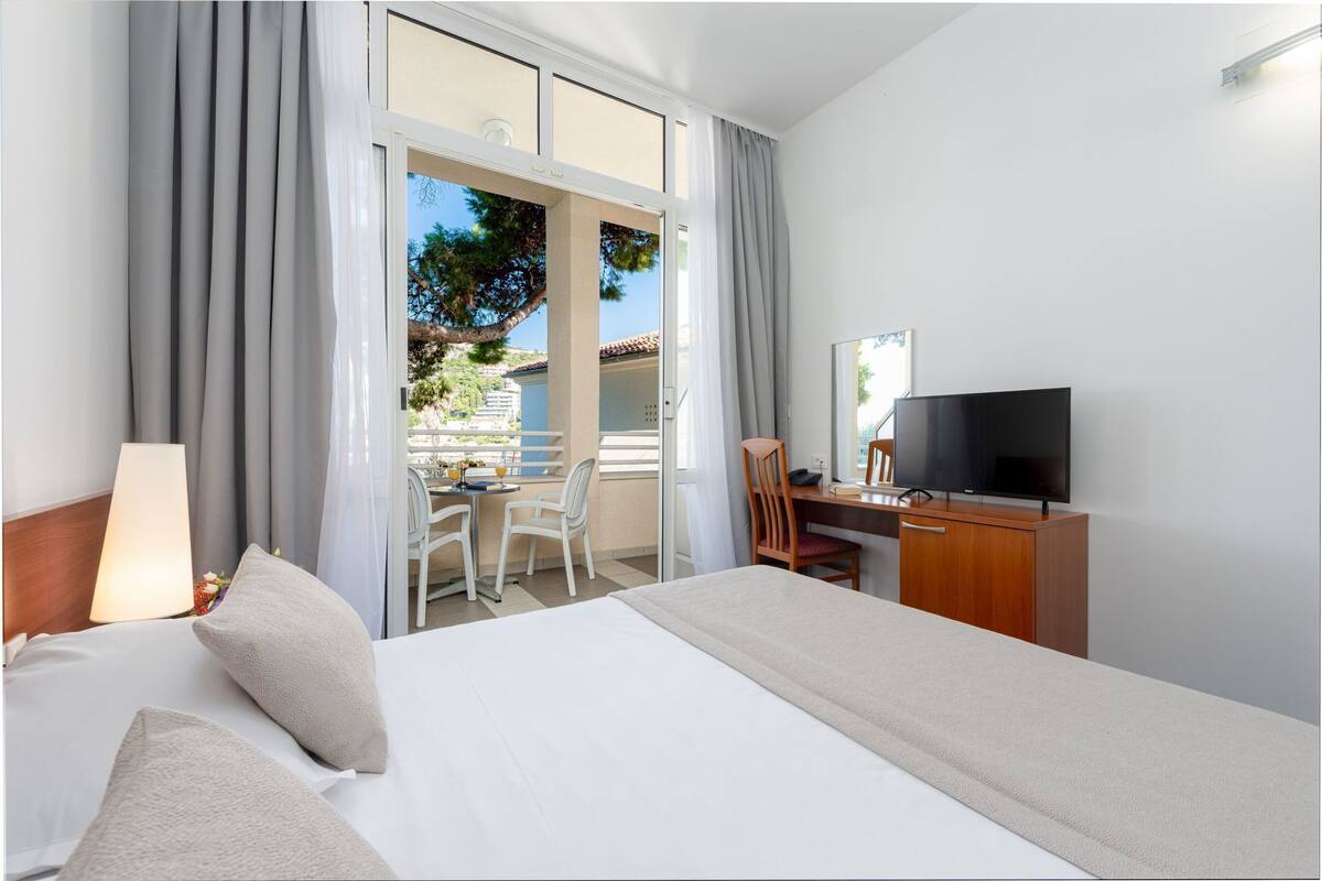 A hotel room with a bed, a desk with a TV, a chair, a lamp, and a view of the outside.
