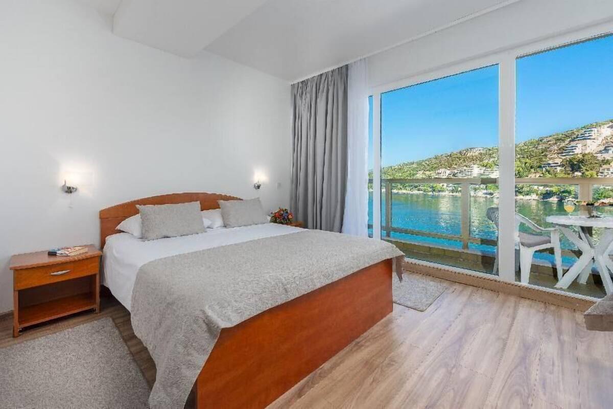 A bedroom with a large bed, a nightstand, a window with a view of a body of water and buildings, and a balcony with a table and chairs.