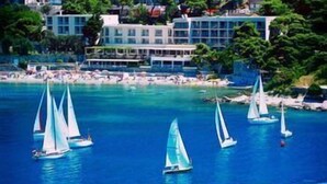 A coastal scene with sailboats on the water, a beach with people, and a building with multiple floors in the background. - Hotel Vis (Dubrovnik)