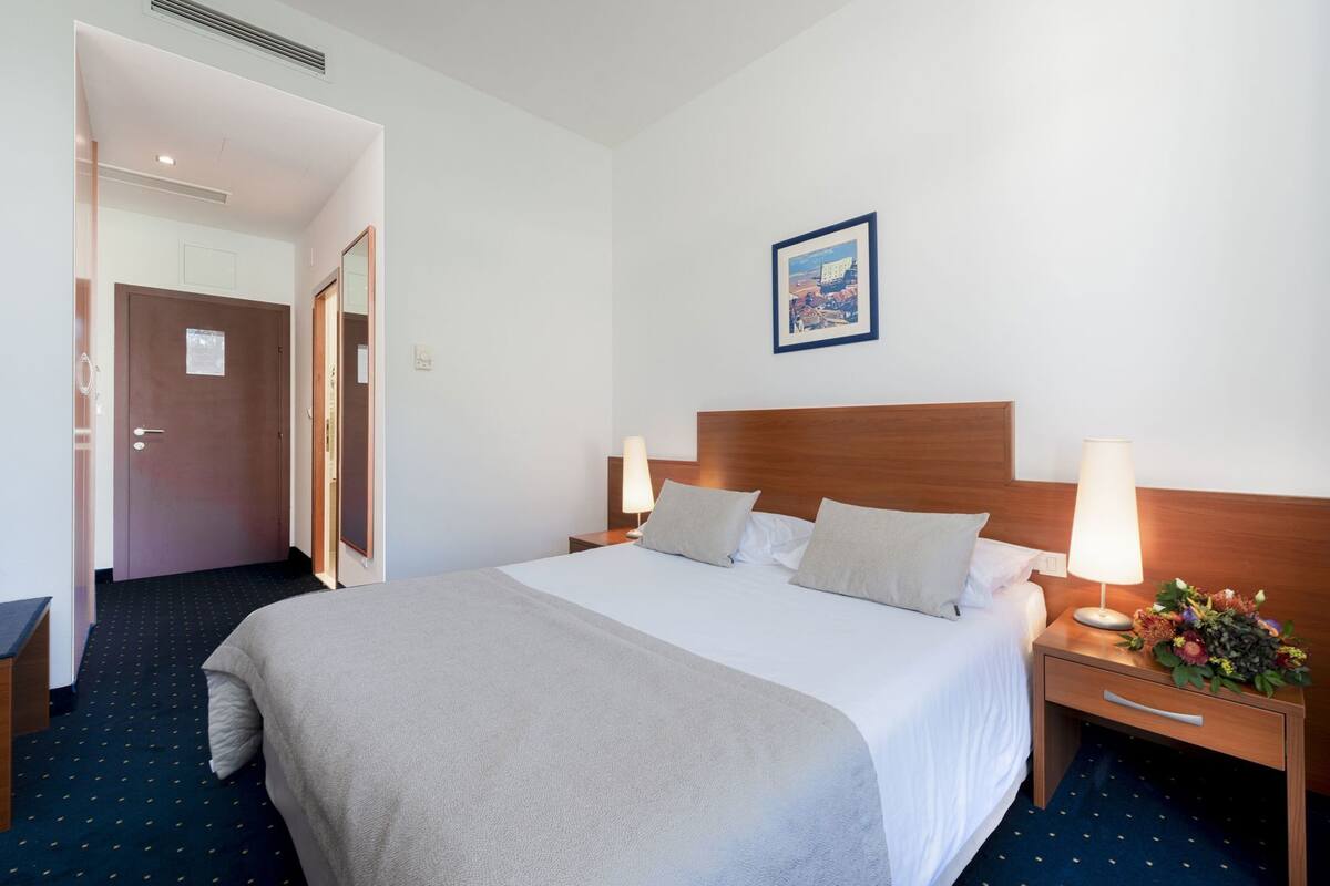 A hotel room with a large bed, two bedside tables with lamps, a wooden headboard, a framed picture on the wall, and a door leading to another room.