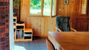 A wooden interior with a dining table, chairs, a bookshelf, and a large window.