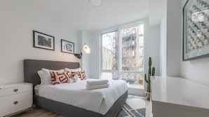 A modern bedroom with a large bed, a nightstand, a lamp, a window with a view of buildings, and a cactus plant. - Stylish 3BR Retreat w Private Deck Near NYC (Hoboken)