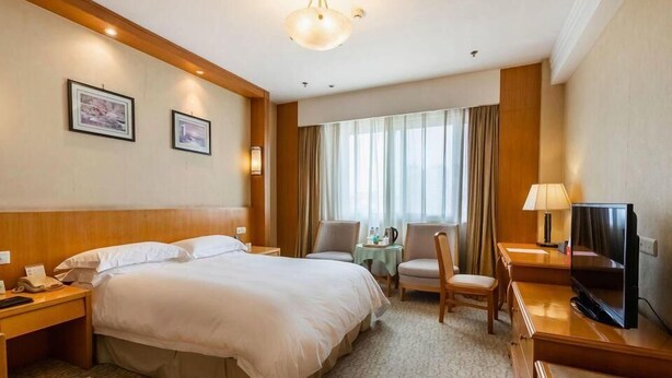 Une chambre d’hôtel avec un grand lit, un bureau avec une télévision, deux chaises, une petite table avec une lampe et une fenêtre avec des rideaux.
