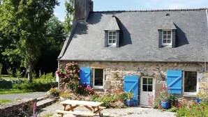 A stone house with a sloped roof, two windows, and blue shutters. There is a wooden picnic table in the front garden.
