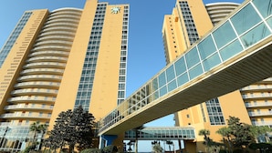 Un moderno edificio de gran altura con fachada curva, un puente peatonal y un cielo azul despejado.