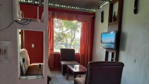 Ein Wohnzimmer mit Fernseher, einer Couch, einem Sessel und einem Tisch. Ein Fenster mit Vorhängen und eine Tür zum Garten.