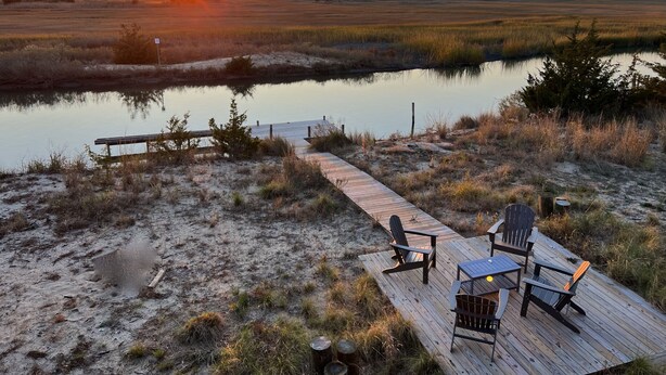 Een houten terras met stoelen en een tafel, met uitzicht op een rustig wateroppervlak en een zonsondergang.