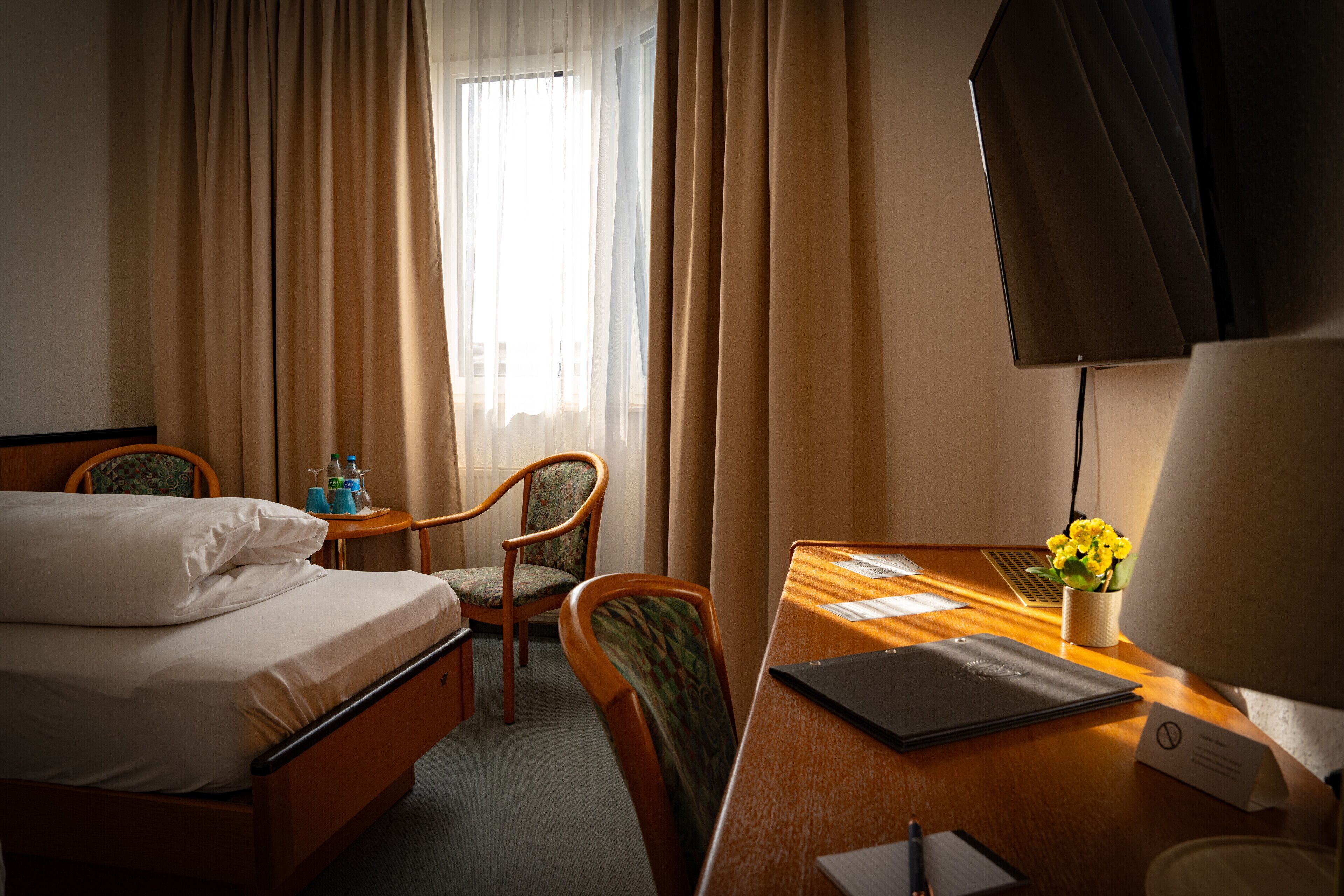 Une chambre d’hôtel avec un lit, un bureau avec un ordinateur portable, une chaise, une petite table avec des bouteilles et une fenêtre avec des rideaux.