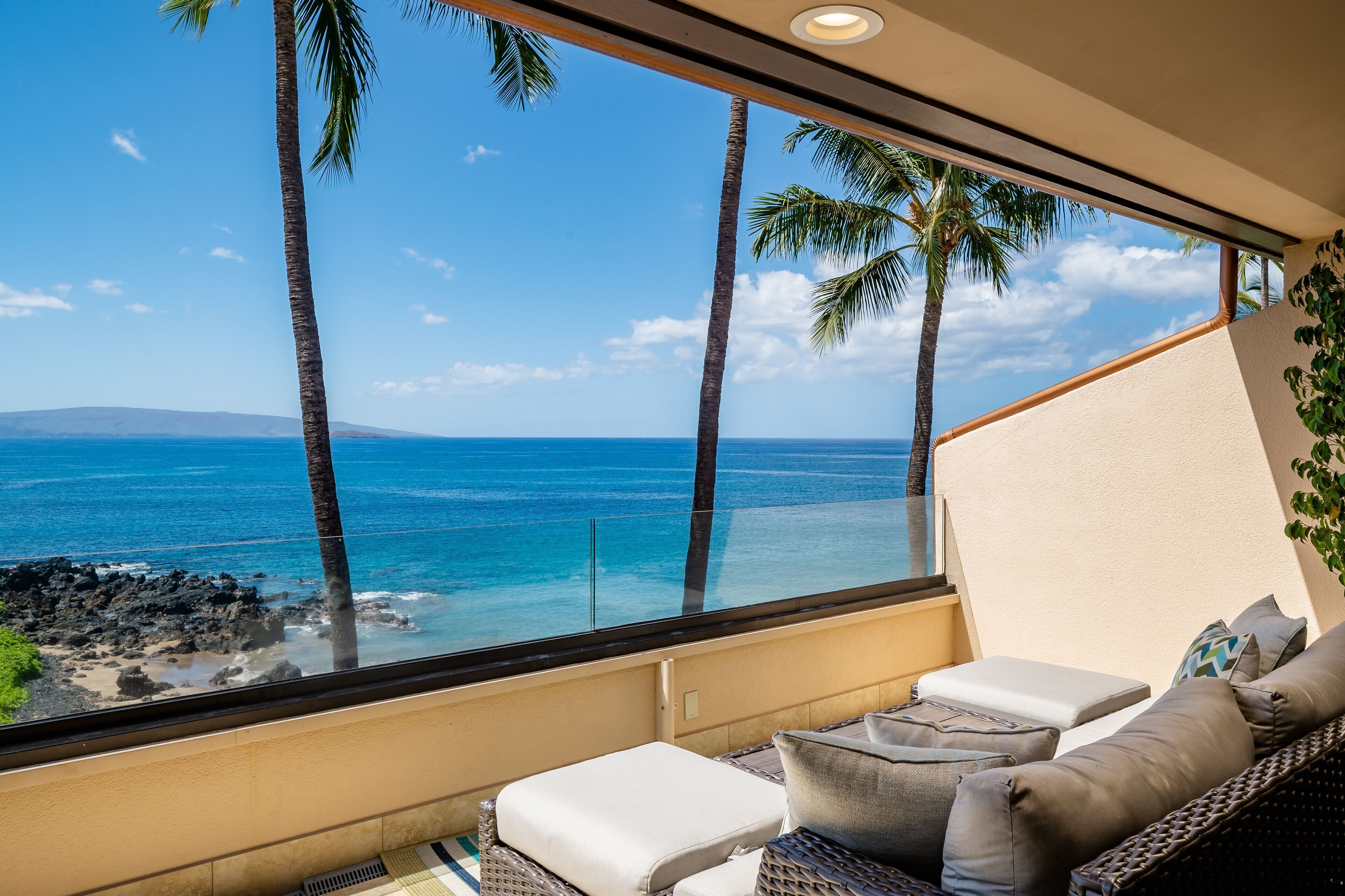 A balcony with a view of the ocean, palm trees, and a wicker sofa with cushions.