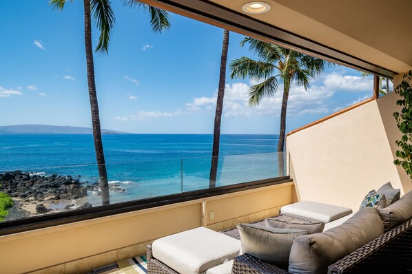 A balcony with a view of the ocean, palm trees, and a wicker sofa with cushions.