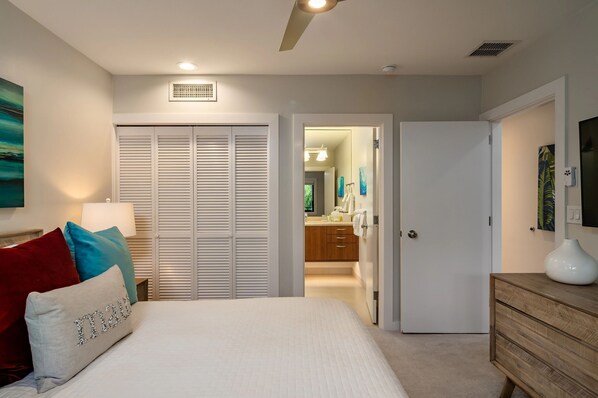 Un dormitorio con cama, mesita de noche, cómoda, armario y baño visible a través de una puerta abierta.