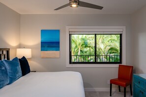 A bedroom with a bed, a nightstand, a lamp, a red chair, a blue cabinet, a ceiling fan, a window with a view of palm trees, and a painting of the ocean. - Expansive One-of-a-Kind Retreat -  3 Oceanview Lanais - Makena Surf G 301-302  (Makena, Maui)
