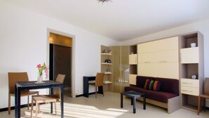 A modern living room with a sofa, a dining table with chairs, and a small coffee table. There is a wardrobe with multiple doors and a shelf unit with various items. - Large studio in Valmy, comfort and calm guaranteed (Lyon)