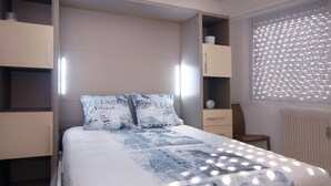 A bedroom with a bed, two pillows, a nightstand, a dresser, and a window with blinds. - Large studio in Valmy, comfort and calm guaranteed (Lyon)