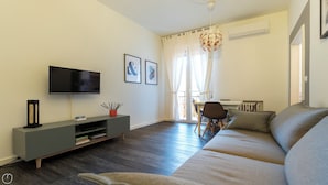 A modern living room with a flat-screen TV, a grey sofa, a dining area with a table and chairs, and a large window with curtains. - Baracca 7 A - One Bedroom Apartment, Sleeps 2 (Bologna)