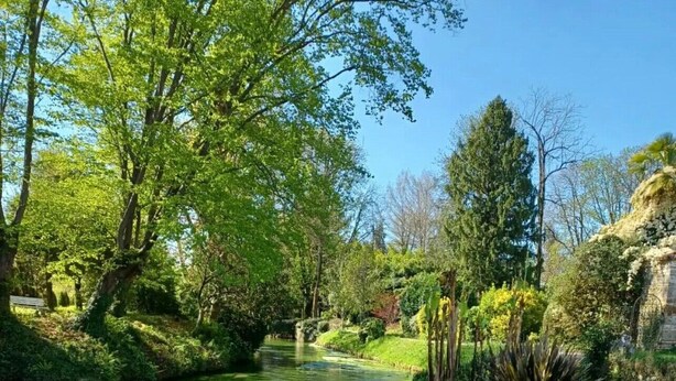 A serene riverside garden with a clear water path, lush greenery, and a stone structure.